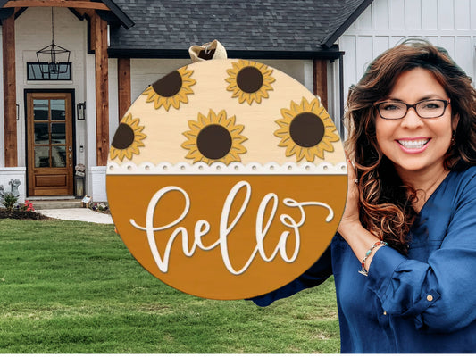 A smiling woman with long brown hair and glasses stands outside a house, holding a round wooden sign decorated with sunflowers and the word “hello” written in white script.