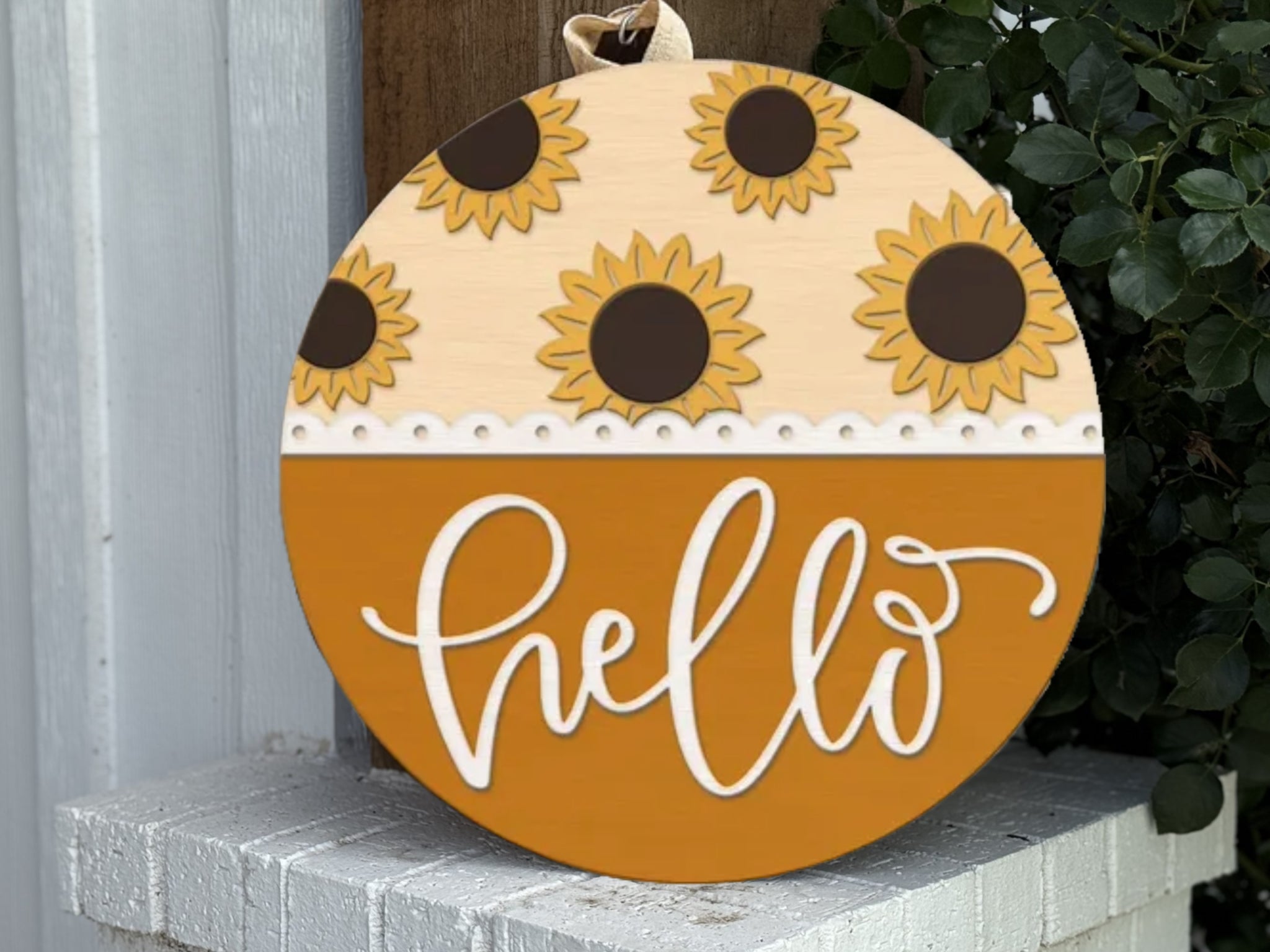 A round wooden sign decorated with sunflowers on the top half and the word hello in white cursive on a mustard yellow background, leaning against a white brick ledge near green leaves.