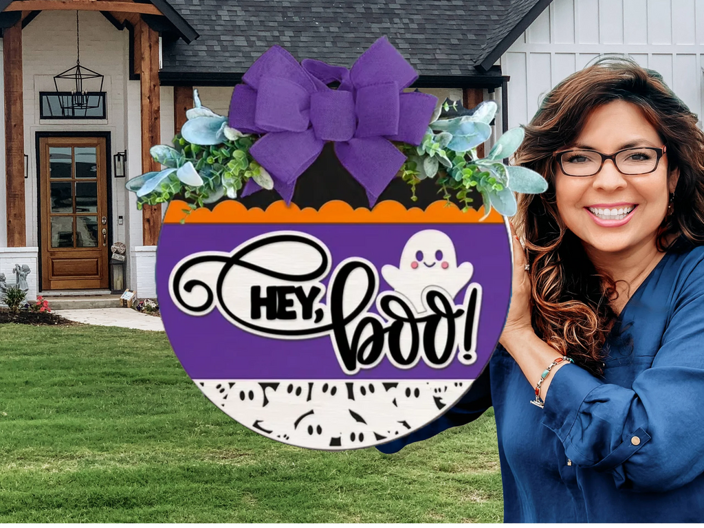 A smiling woman with wavy hair and glasses stands outside a house, holding a round Halloween sign with a purple bow, leaves, a ghost, and the text HEY, BOO! in front of the door.