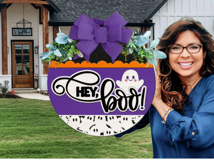 A smiling woman with wavy hair and glasses stands outside a house, holding a round Halloween sign with a purple bow, leaves, a ghost, and the text HEY, BOO! in front of the door.