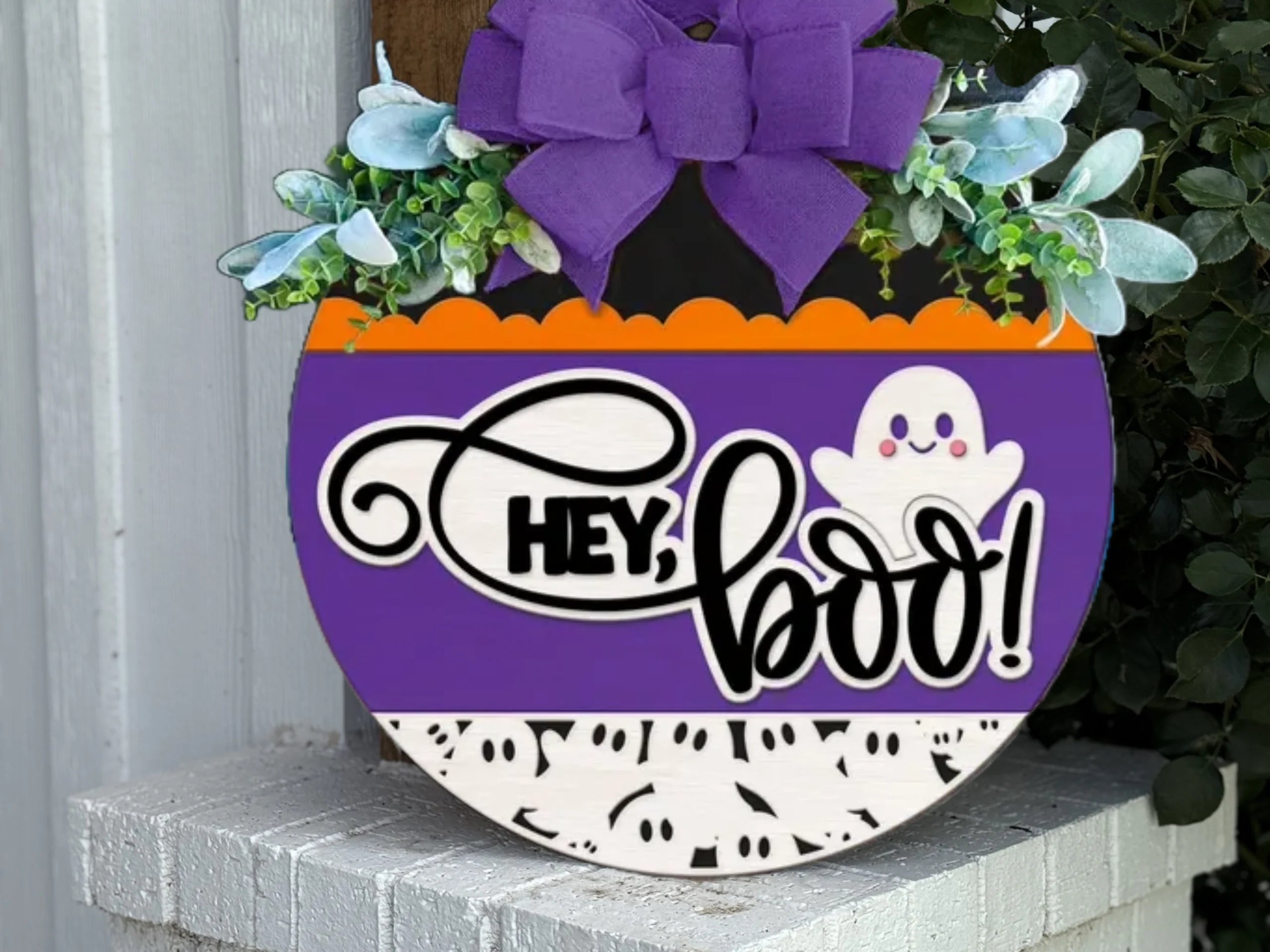 A round Halloween sign with a purple bow, greenery, and the words Hey, boo! next to a smiling ghost. The sign has purple, orange, and white sections with black ghost and dot patterns.