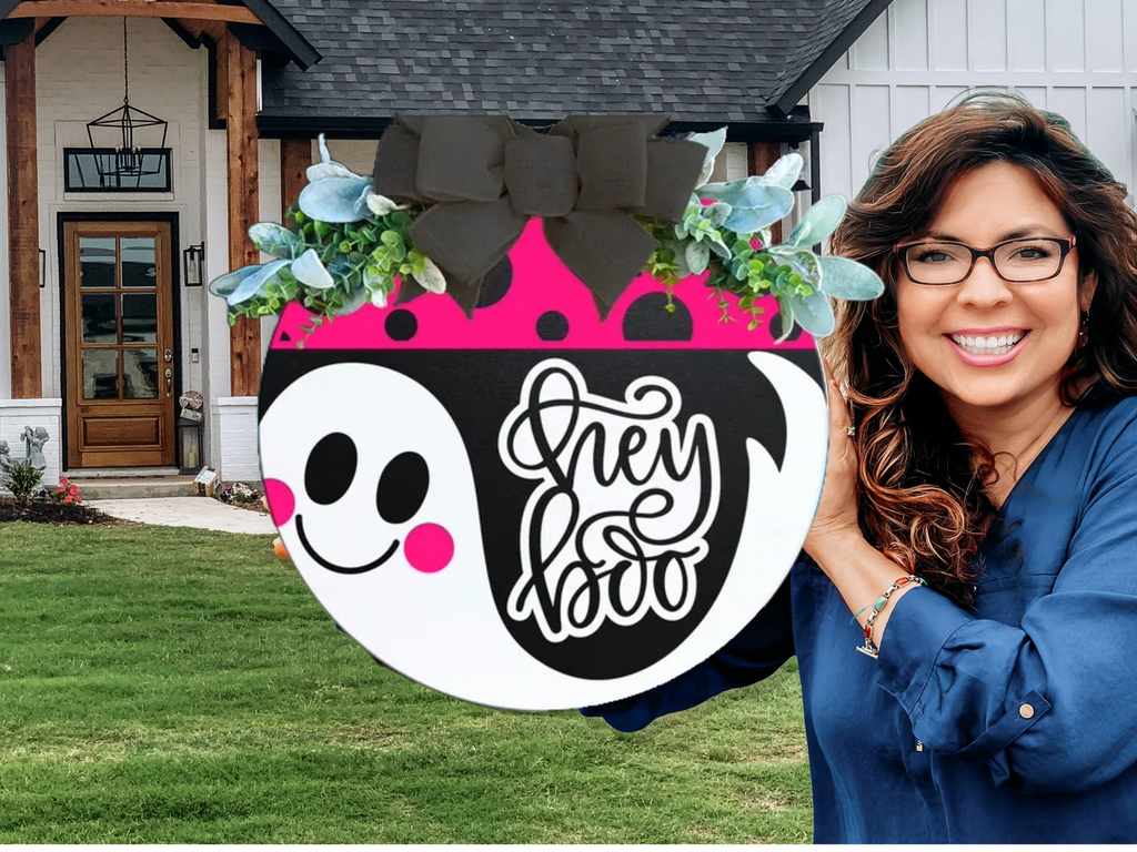 A smiling woman with wavy brown hair and glasses holds a round decorative sign featuring a cute ghost, plants, a big black bow, and the words Hey Boo in front of a modern house.