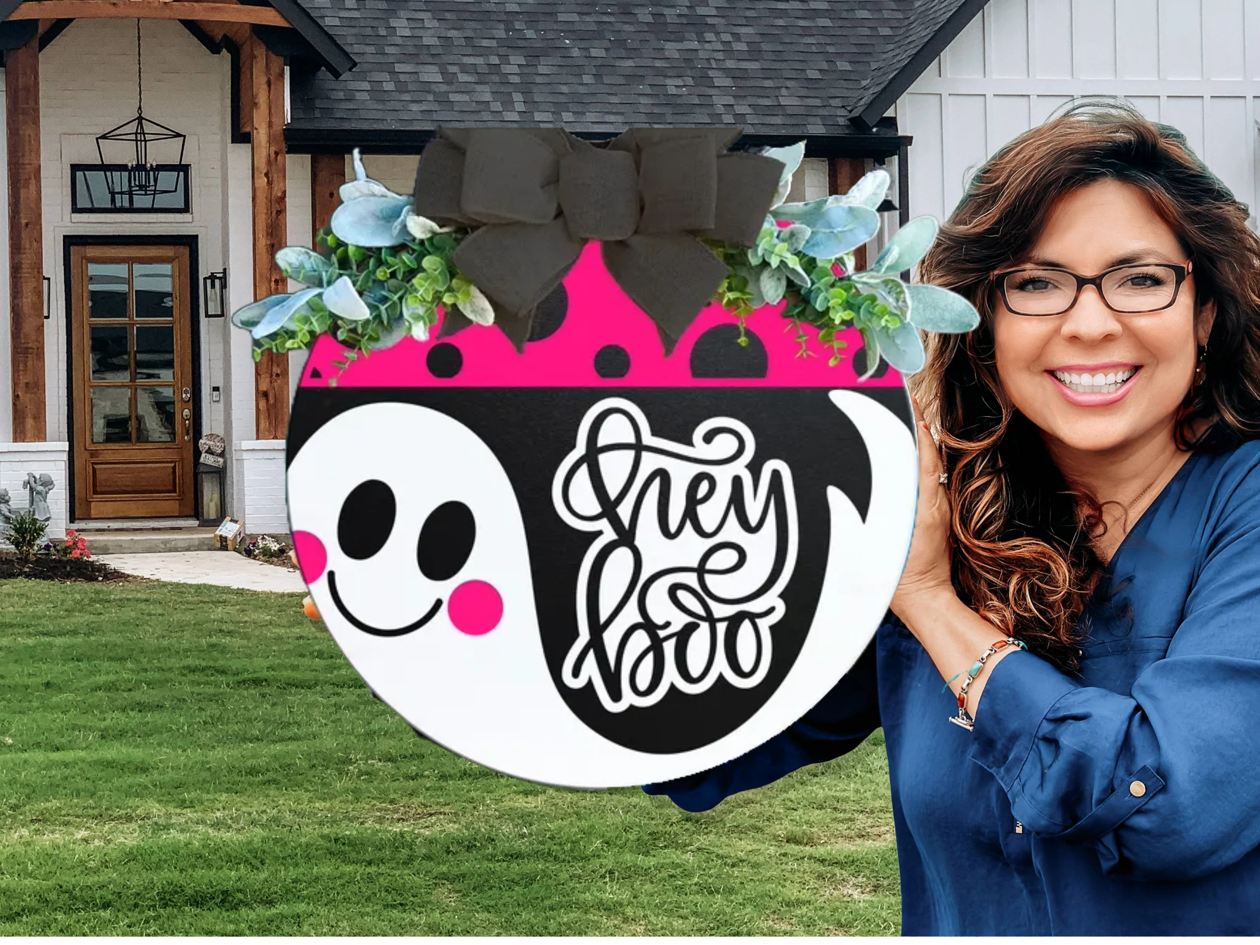 A smiling woman with wavy brown hair and glasses holds a round decorative sign featuring a cute ghost, plants, a big black bow, and the words Hey Boo in front of a modern house.