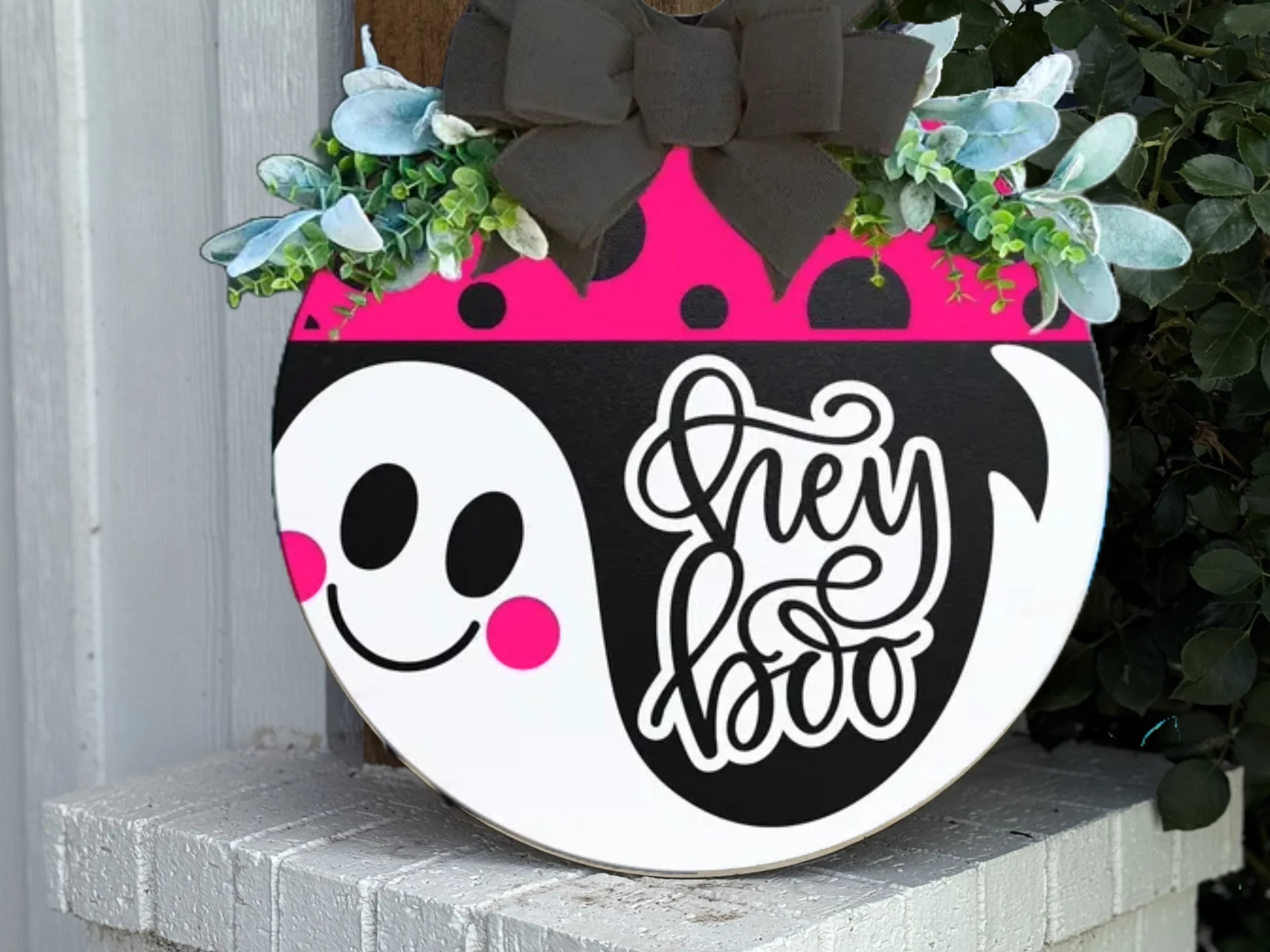 A round sign with a pink and black top, green leaves, and a black bow. The sign features a smiling ghost with pink cheeks and the words hey boo in white script on a black background.