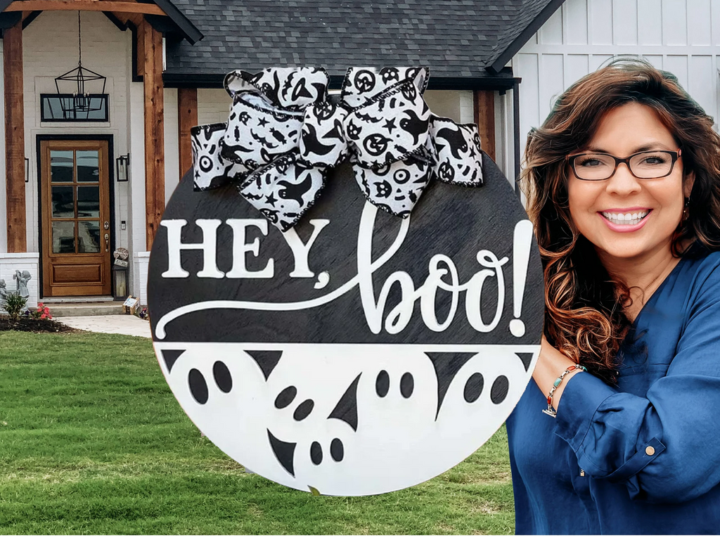 A smiling woman with glasses holds a round sign decorated with a large black-and-white bow, ghost patterns, and the words HEY, boo! with a house and green lawn in the background.