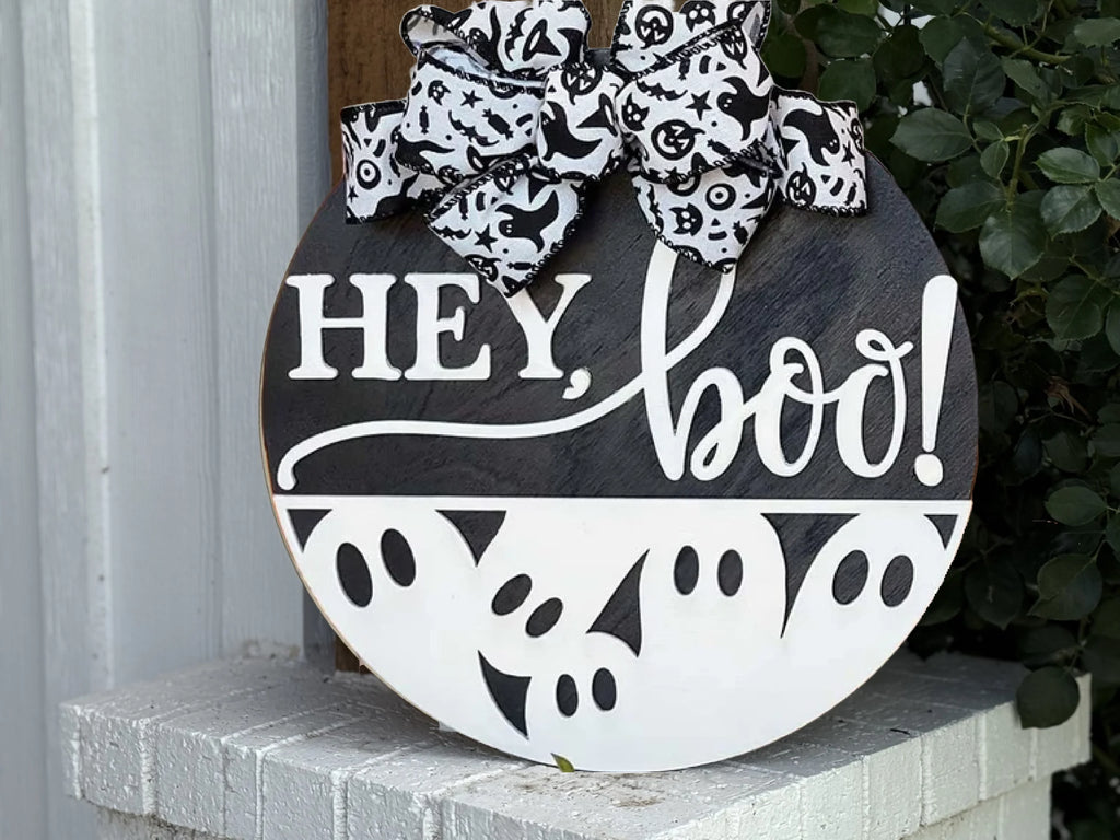 A round wooden sign with HEY, boo! written in white, featuring ghost faces along the bottom and topped with a black-and-white Halloween-themed bow, hangs on a white brick ledge near green leaves.