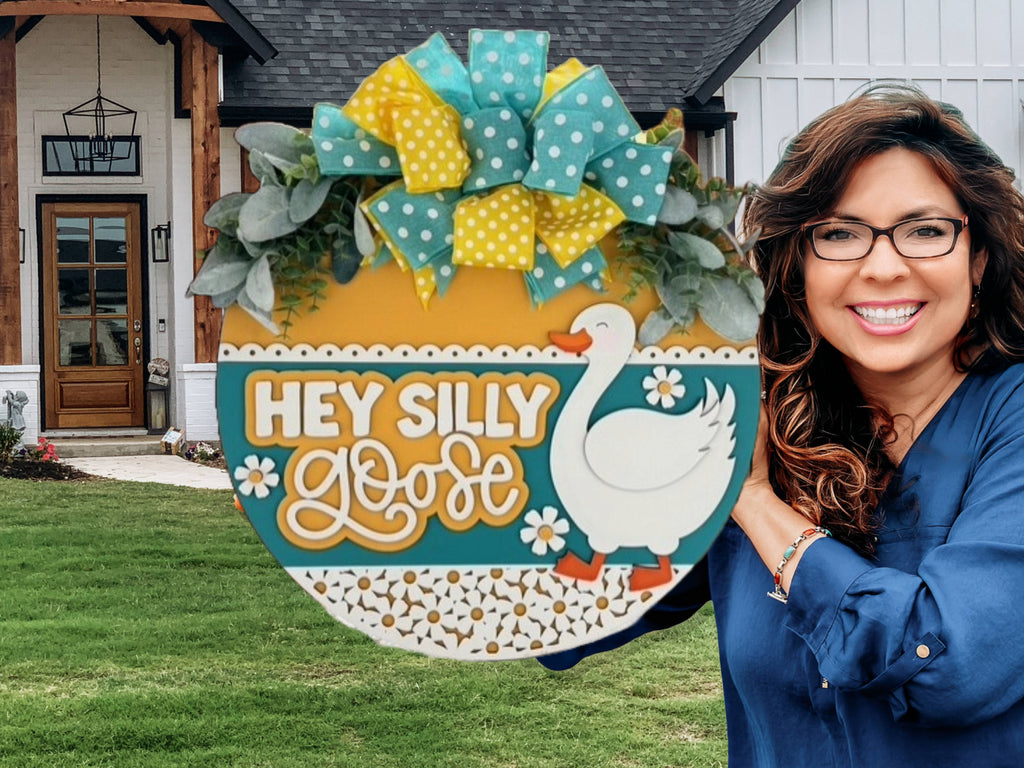 A smiling woman holding a round sign decorated with a goose, yellow and blue bows, and the text HEY SILLY GOOSE stands outside a modern house with a lawn and wooden front porch.