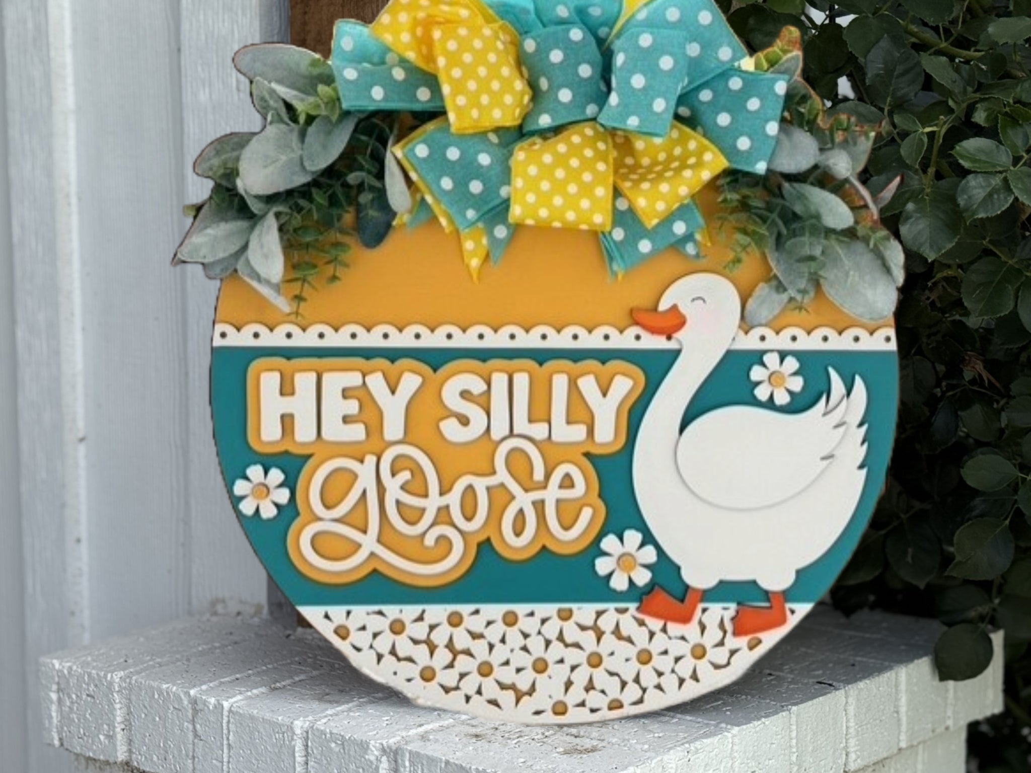 A decorative sign with a white goose, yellow and teal patterns, greenery, and a yellow-and-teal polka dot bow. The sign reads HEY SILLY GOOSE in bold letters.