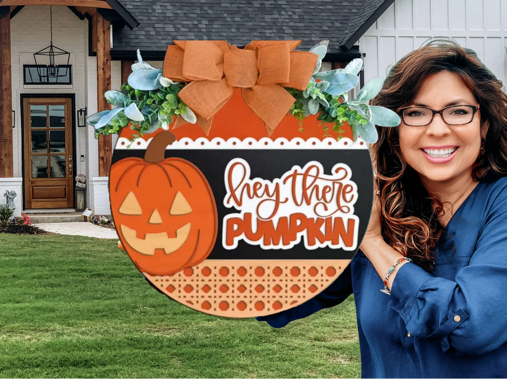 A smiling woman holds a round Halloween sign decorated with a bow, greenery, and a pumpkin. The sign reads “hey there pumpkin.” She stands outside in front of a modern house with a green lawn.