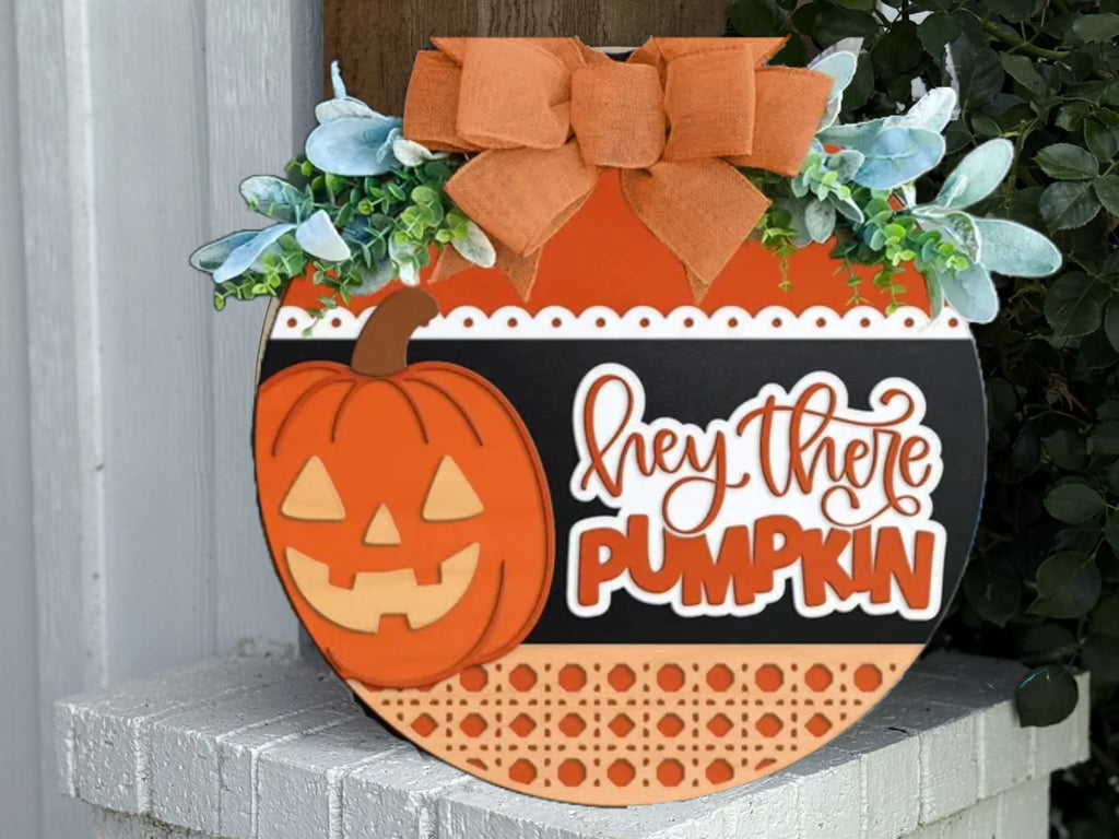 A round, decorative Halloween sign features a smiling jack-o-lantern, greenery, an orange bow, and the words hey there PUMPKIN in playful lettering on a white, black, and orange background.
