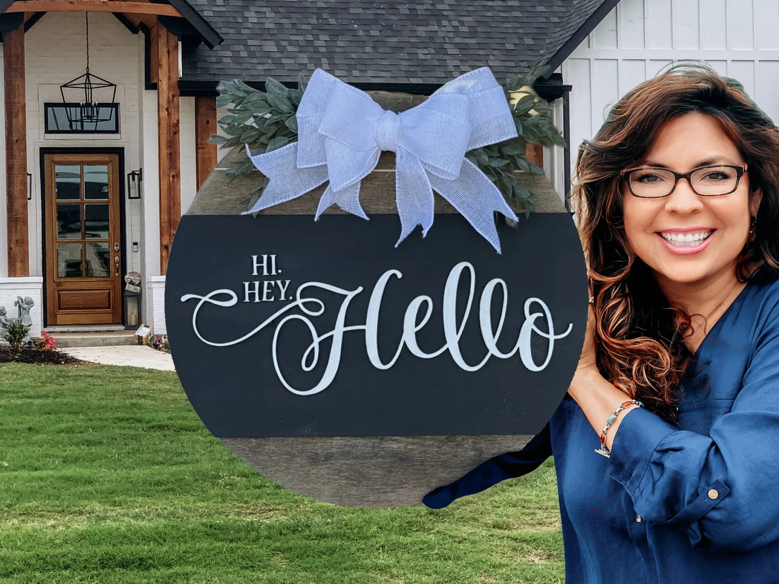 A smiling woman with long brown hair and glasses holds the Welcome Door Sign, Welcome Door Wreath, Hi Hey Hello—a round black sign with a blue bow—in front of a modern house with a green lawn.