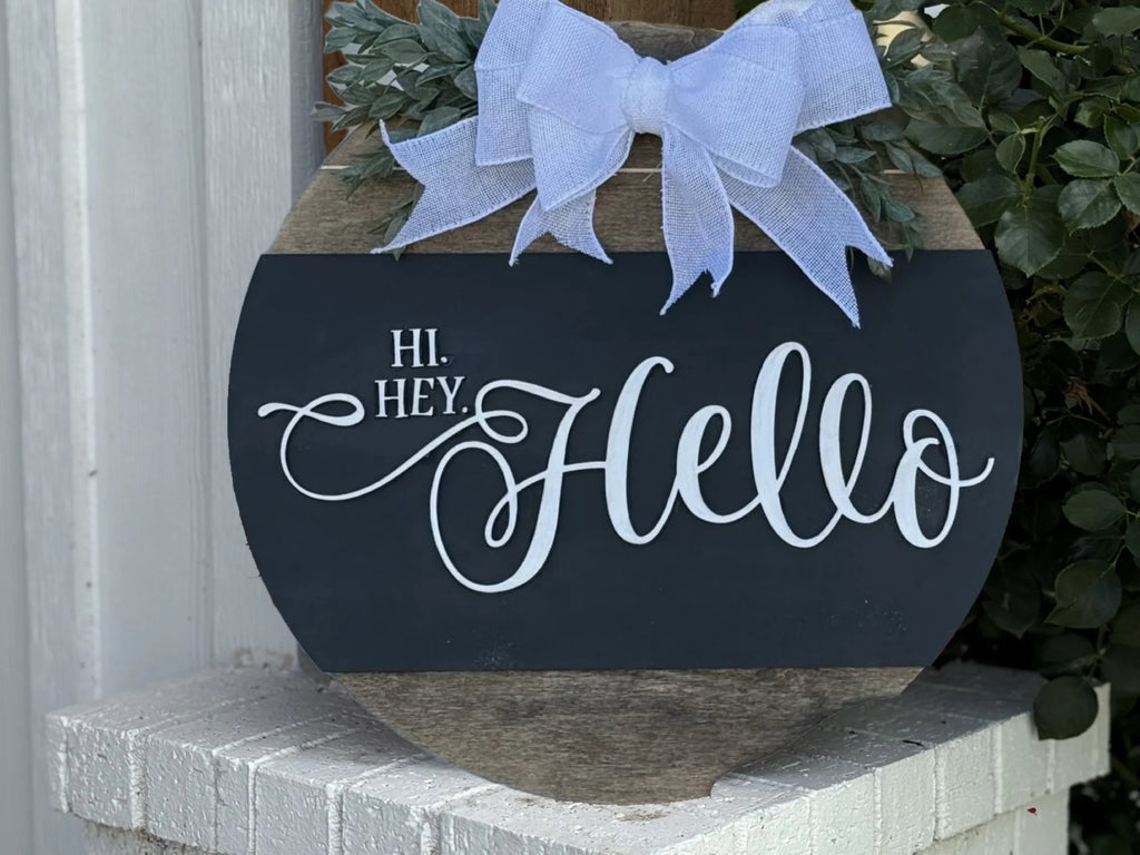 The Welcome Door Sign, Welcome Door Wreath, Hi Hey Hello features a round wooden design with a large light blue bow, green leaves, and Hi. Hey! Hello in white script, displayed against a white brick surface with greenery nearby.
