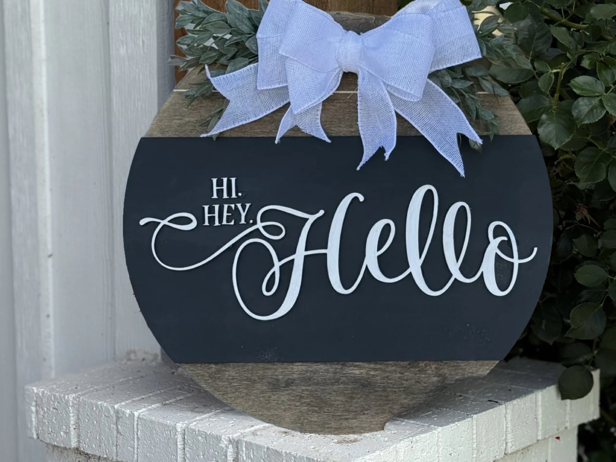 The Welcome Door Sign, Welcome Door Wreath, Hi Hey Hello features a round wooden design with a large light blue bow, green leaves, and Hi. Hey! Hello in white script, displayed against a white brick surface with greenery nearby.