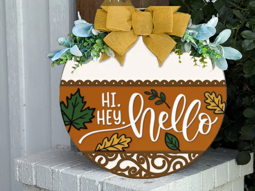 A round wooden sign decorated with green leaves, a yellow bow, and autumn motifs sits on a white brick step. The sign reads Hi. Hey. Hello with leaf illustrations and elegant swirl designs.