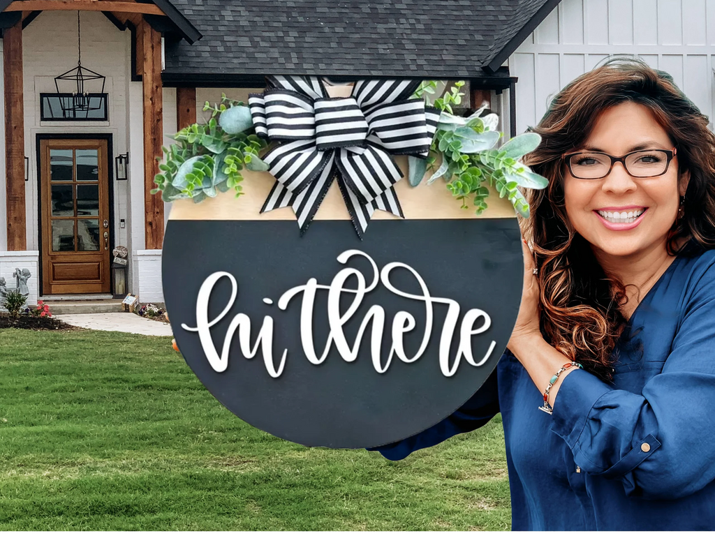A smiling woman with long brown hair and glasses holds a round black sign with hi there in cursive white letters, decorated with greenery and a striped bow, standing in front of a modern house.