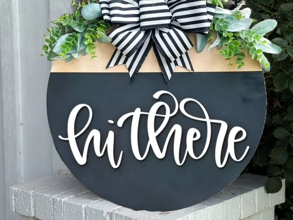 A round black sign with white script reading hi there, decorated with greenery and a black-and-white striped bow at the top, displayed outdoors.