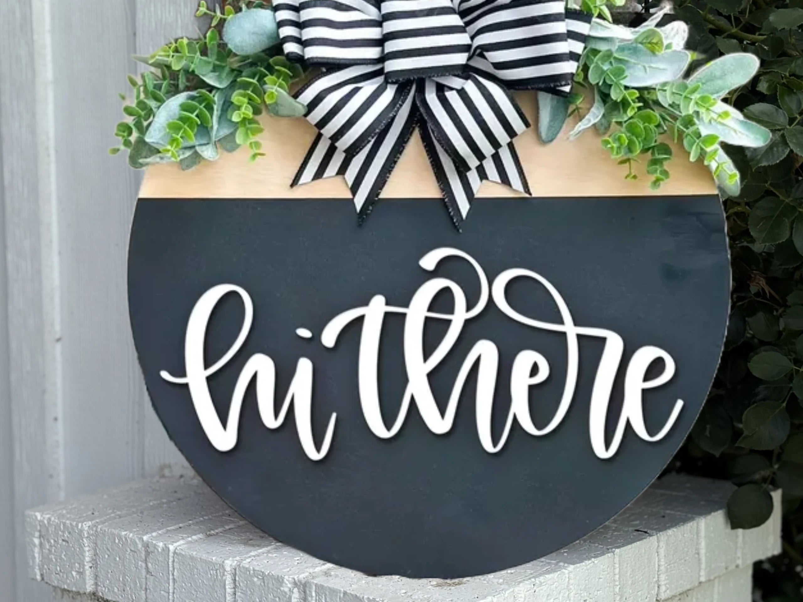 A round black sign with white script reading hi there, decorated with greenery and a black-and-white striped bow at the top, displayed outdoors.