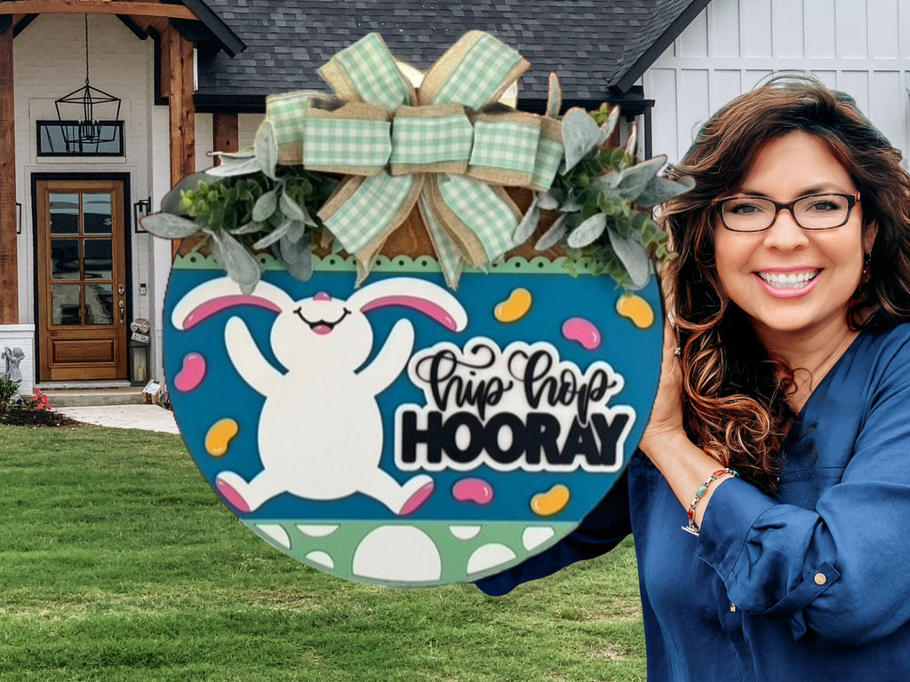 A smiling woman with wavy brown hair and glasses holds a colorful Easter-themed sign shaped like an egg, featuring a bunny, jellybeans, greenery, and the text “Hip Hop HOORAY,” in front of a modern house with a lawn.