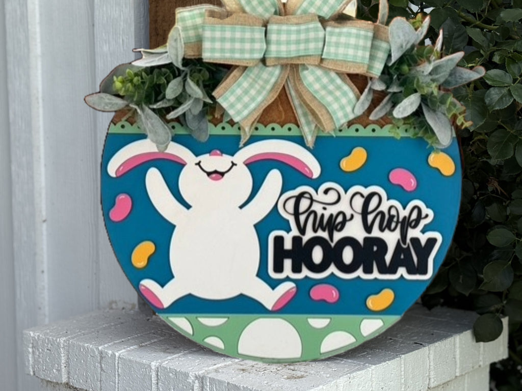A decorative sign features a cheerful white bunny with pink ears, surrounded by colorful jelly beans. Above the bunny is greenery and a green plaid bow; below is text reading Hip Hop HOORAY. The sign rests on a white brick surface.