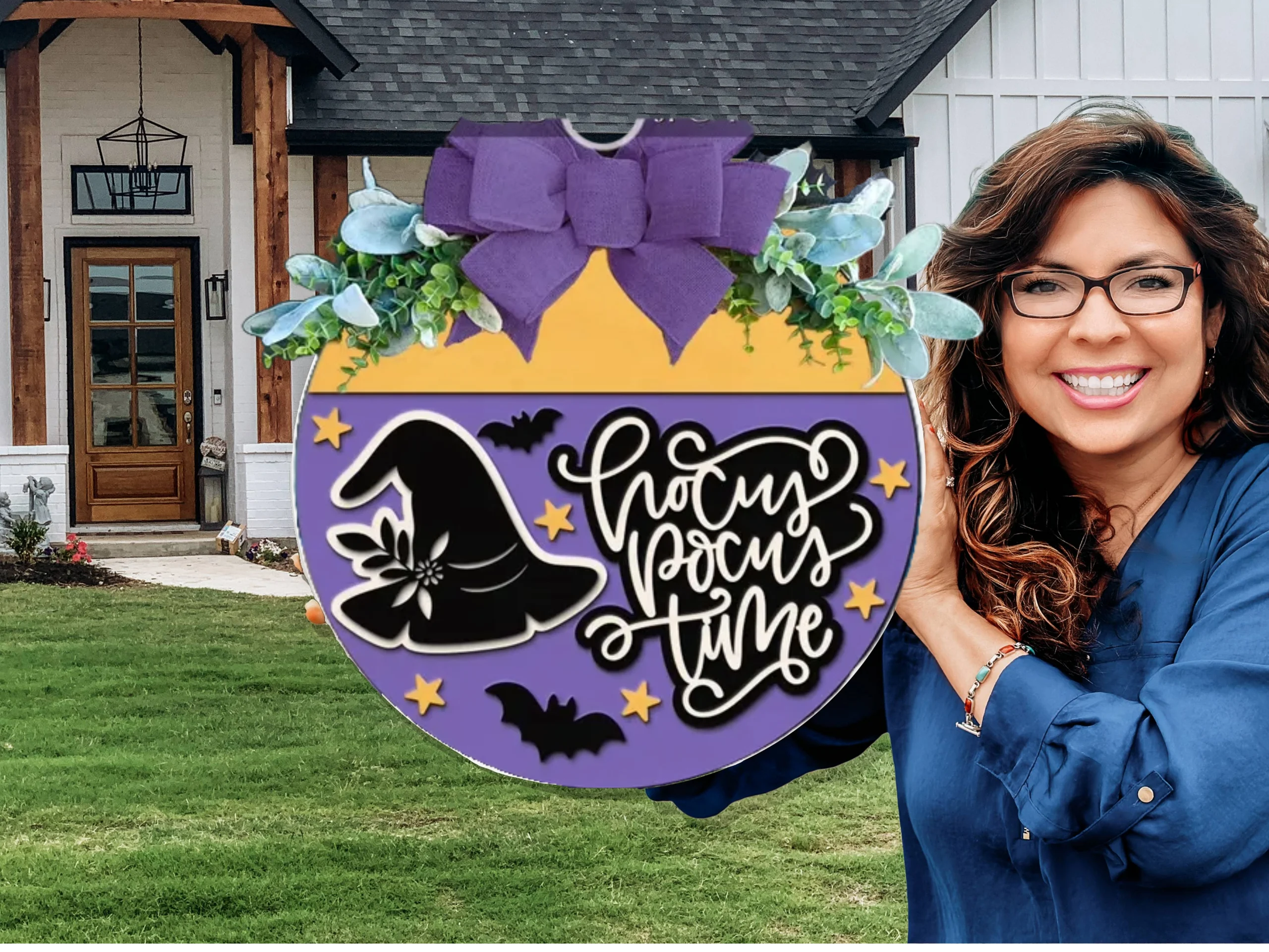 A smiling woman holds a round, purple and yellow Halloween sign with a witch hat, bats, stars, greenery, and a large purple bow. The sign reads Hocus Pocus Time. A house with a lawn is in the background.