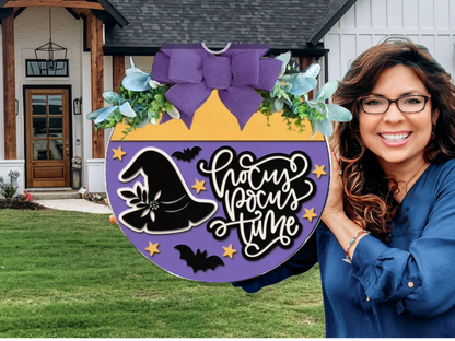 A smiling woman holds a round, purple and yellow Halloween sign with a witch hat, bats, stars, greenery, and a large purple bow. The sign reads Hocus Pocus Time. A house with a lawn is in the background.