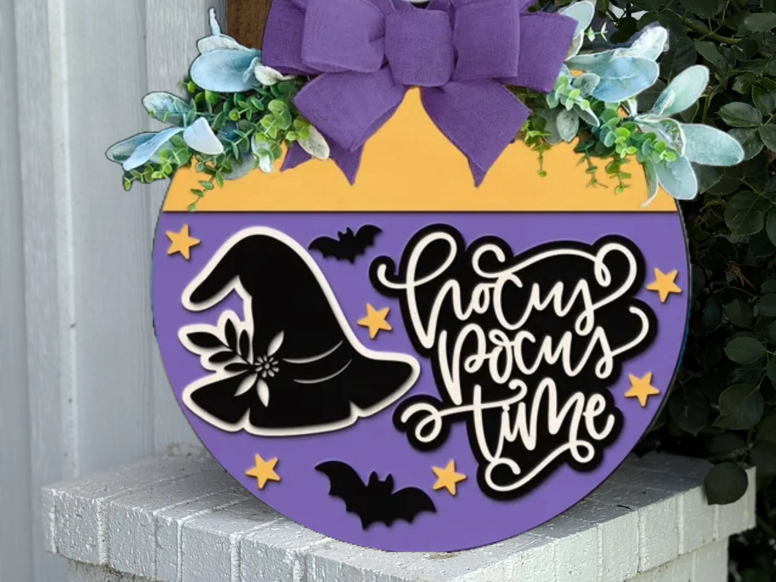 A round Halloween sign with a purple bow, greenery, and blue flowers on top. The sign reads Hocus Pocus Time and features a witch hat, bats, and yellow stars on a purple and yellow background.