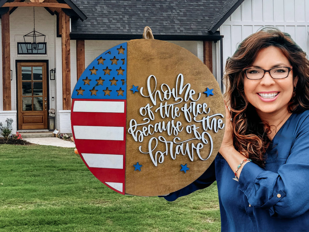 A smiling woman stands outside a house, holding a large round sign with an American flag design and the words Home of the free because of the brave in script. The house and yard are visible in the background.
