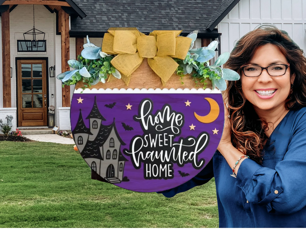 A smiling woman stands outside a house, holding a round sign decorated with greenery and a large yellow bow. The sign reads, Home Sweet Haunted Home with a spooky house, bats, moon, and stars on a purple background.
