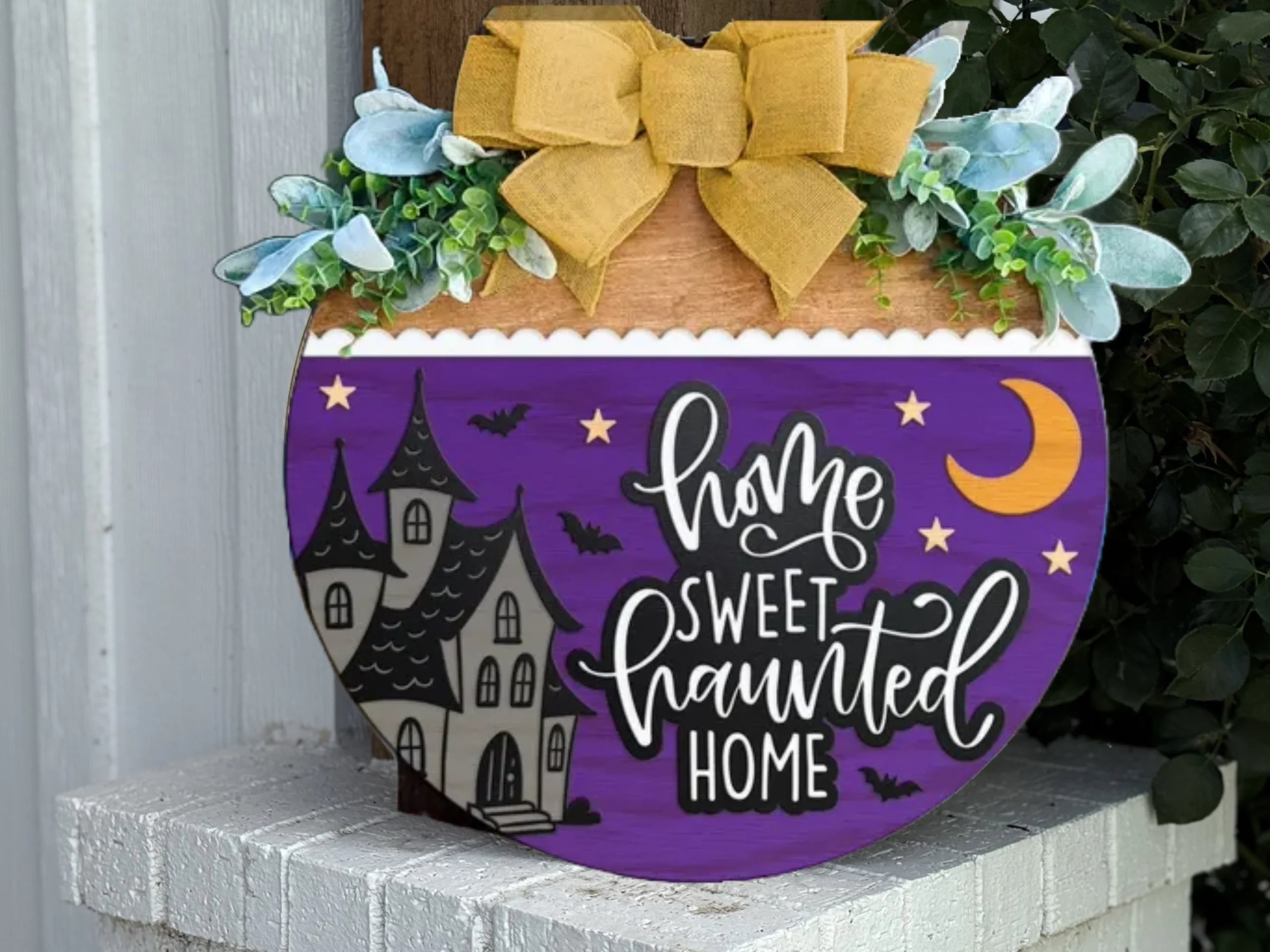 A round purple sign with a haunted house, bats, and a crescent moon, reads Home Sweet Haunted Home. It is topped with green leaves and a large yellow bow, and is displayed on a white brick ledge.