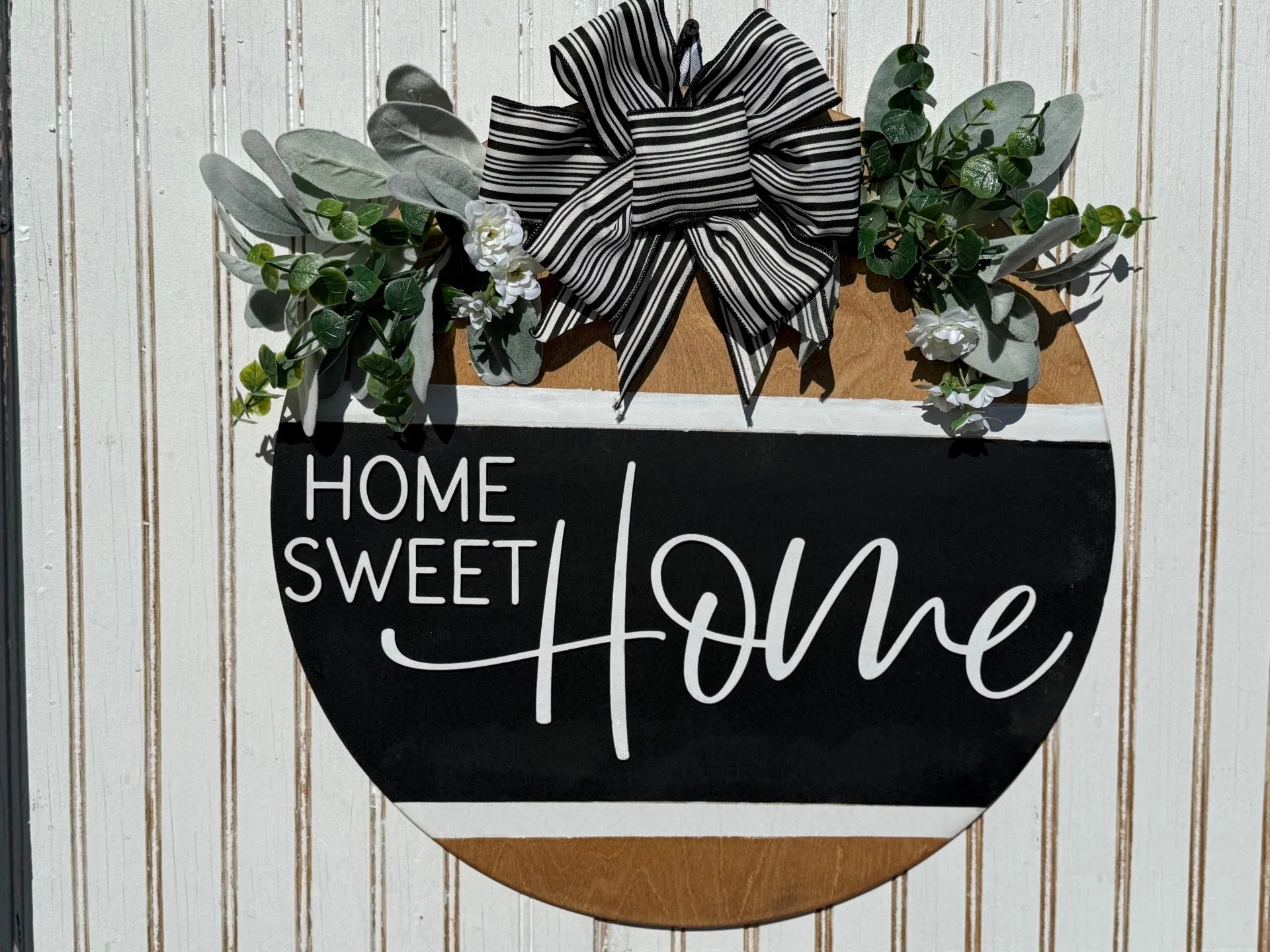 The Welcome To Our Home door sign is a round wooden piece adorned with a black and white Home Sweet Home script. It features a large striped bow, green leaves, and small white flowers against a textured white wood background—ideal for welcoming guests.