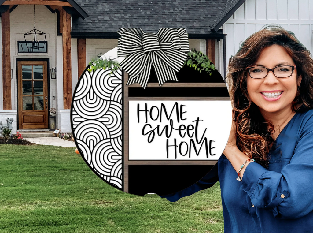 A smiling woman with wavy hair and glasses holds a round sign that reads Home Sweet Home in front of a modern house with white walls, wood accents, and a green lawn.