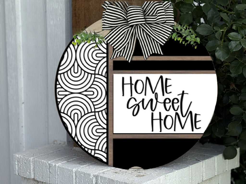 A round decorative sign with a black and white bow and greenery on top. The sign features black and white geometric patterns on one side and the text Home Sweet Home in a playful font on the other.