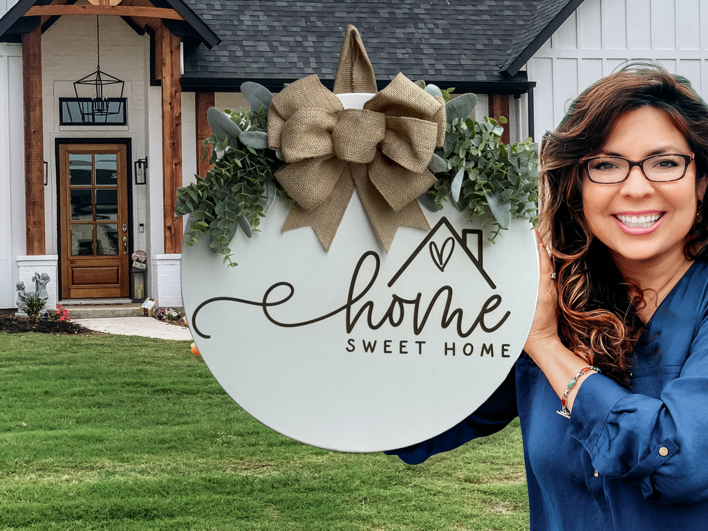 A smiling woman in glasses holds a decorative round sign with greenery and a bow that reads Home Sweet Home, standing in front of a modern house with a wooden front door and green lawn.