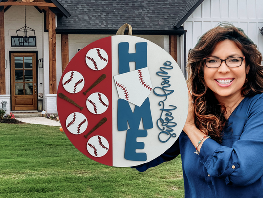 A smiling woman with long brown hair and glasses holds a round sign decorated with baseballs, bats, the word HOME, and Sweet Game, standing in front of a white house with a wooden porch.