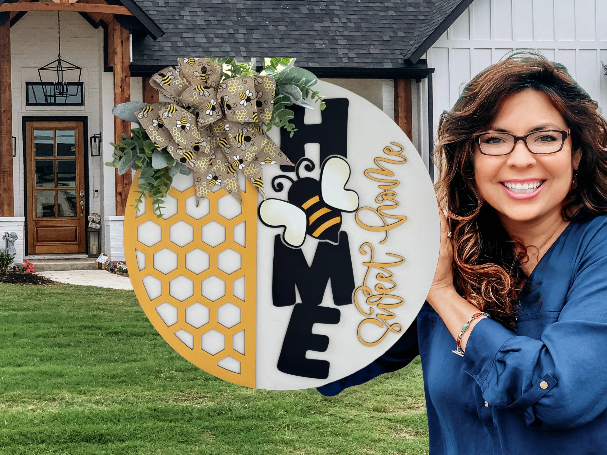Woman in a blue shirt holding a round decorative sign outside a house. The sign features a bee, honeycomb pattern, and the word HOME, with Sweet Home written in cursive. A floral and ribbon arrangement is at the top. The house has a wooden door and white siding.