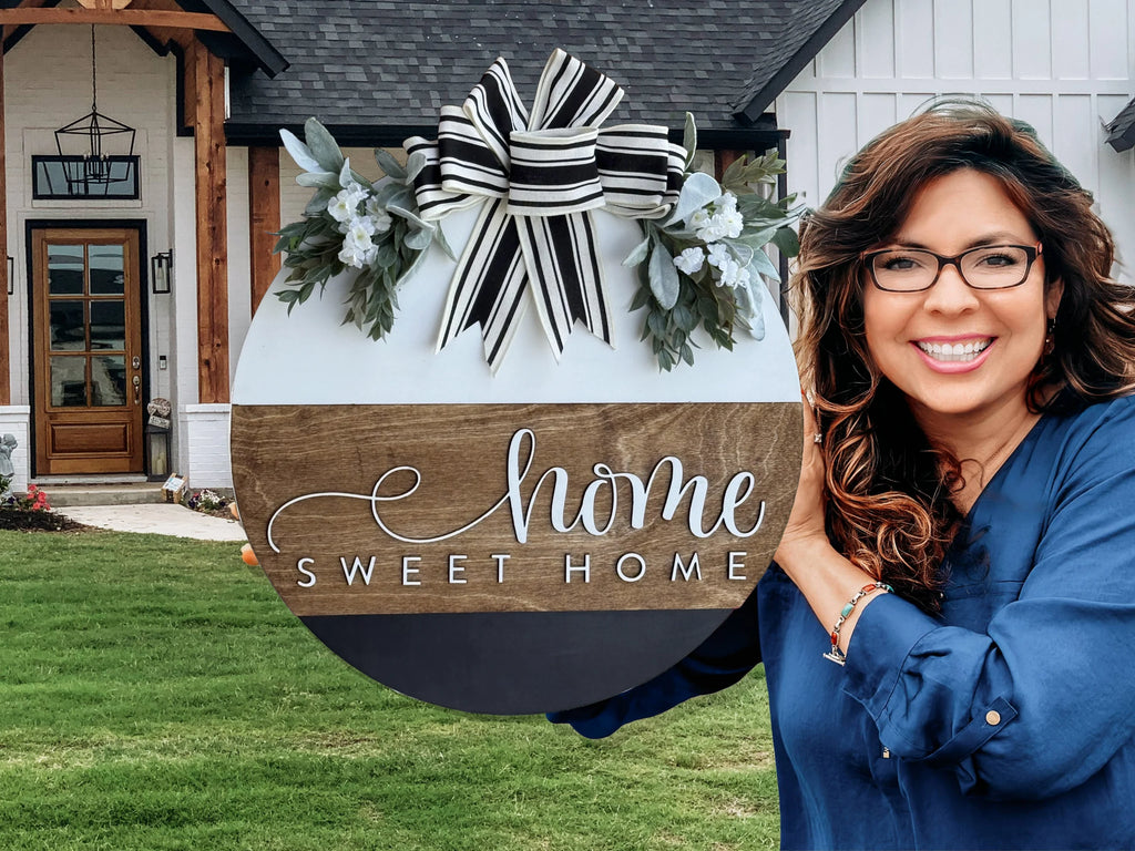 A smiling woman with wavy hair and glasses holds a round Home Sweet Home sign decorated with greenery and a striped bow, standing in front of a modern farmhouse-style home.