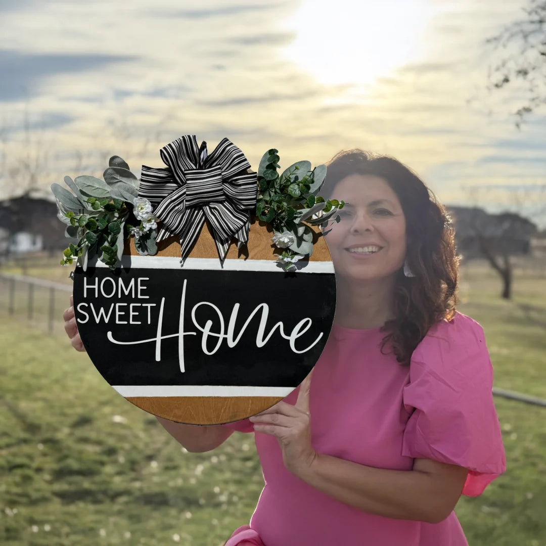 A woman in a pink dress stands in grass, holding the Welcome To Our Home door sign. The round wreath has Home Sweet Home in white script on a black background, adorned with leaves and a striped ribbon. Soft sunlight filters through clouds, casting warmth on the charming display.