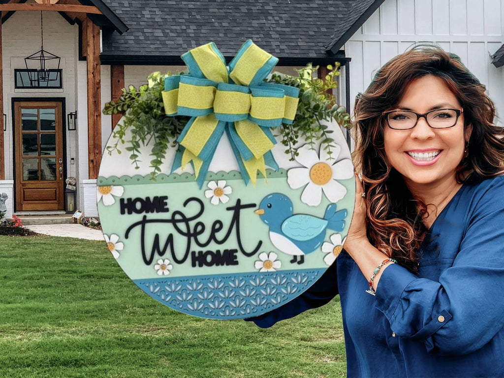 Smiling woman holding a circular sign that reads Home Tweet Home with a blue bird and daisies. The sign has a large, decorative bow and greenery on top. Shes standing in front of a modern house with a wooden door and lantern. Lush grass is in the foreground.