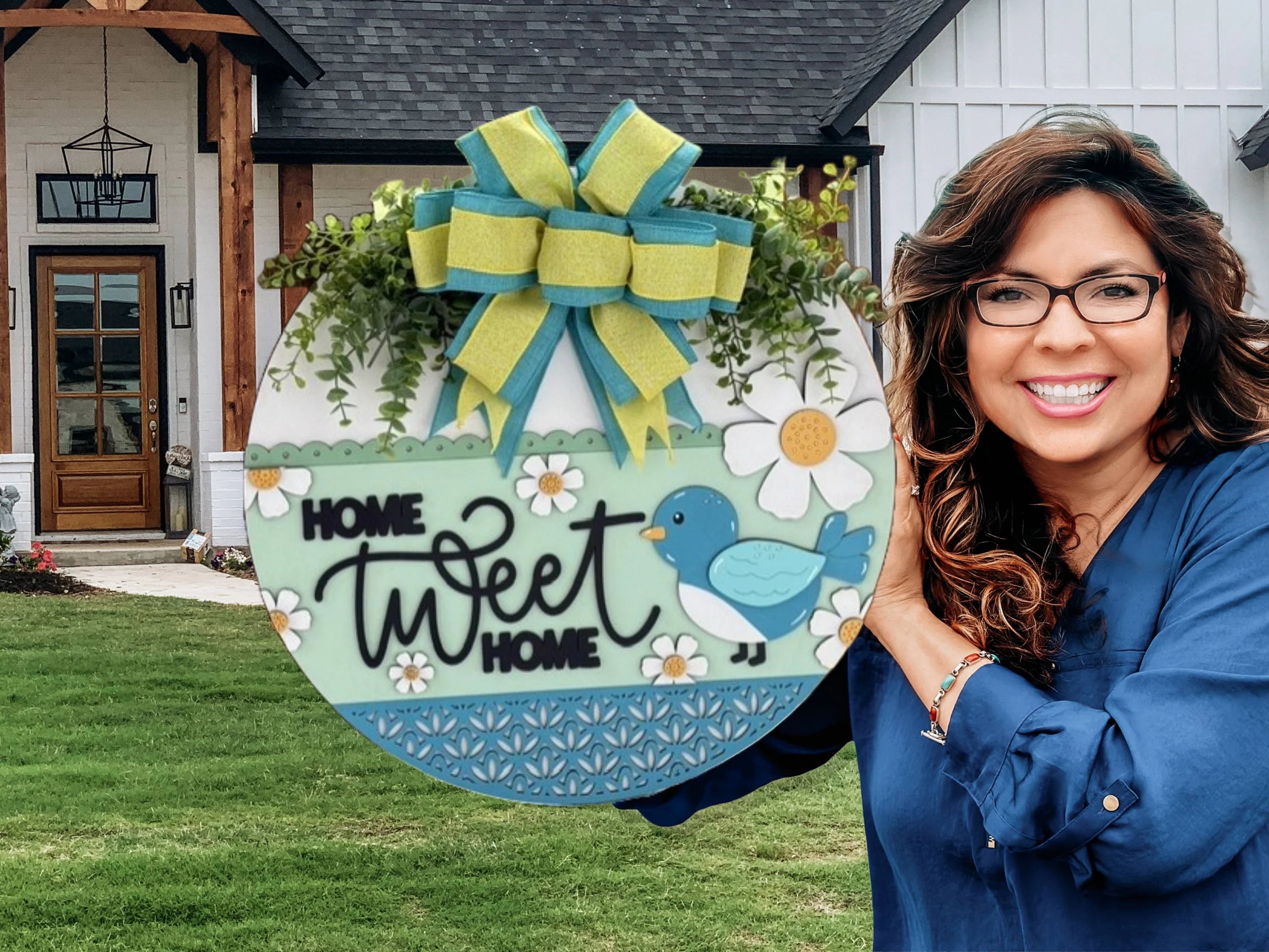 Smiling woman holding a circular sign that reads Home Tweet Home with a blue bird and daisies. The sign has a large, decorative bow and greenery on top. Shes standing in front of a modern house with a wooden door and lantern. Lush grass is in the foreground.