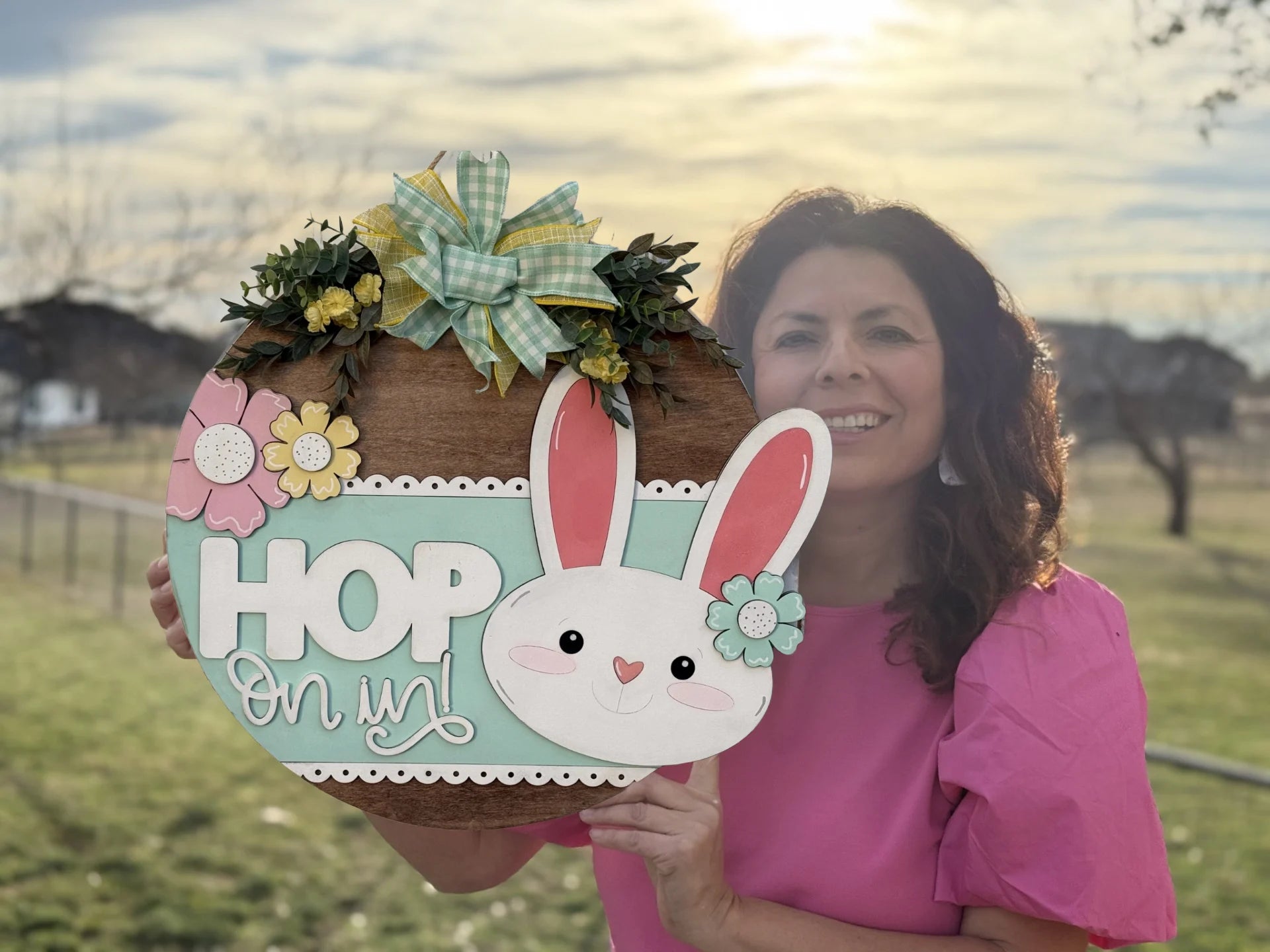 A woman in a pink blouse stands outdoors, smiling and holding a round wooden Easter sign. The sign features a white bunny face with pink ears, the words HOP On in!, and is decorated with flowers and ribbon. The background shows a grassy field and a cloudy sky.