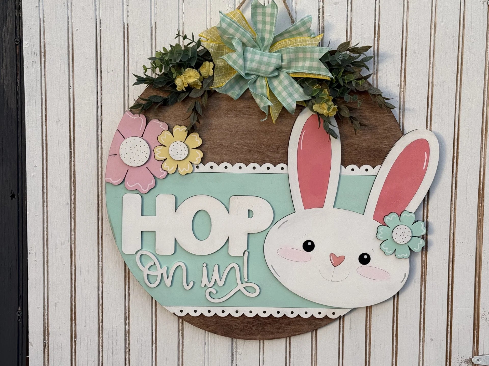 The Hop On In Door Hanger is a round wooden sign featuring an Easter bunny face and the phrase Hop on in! Its decorated with colorful flowers, a green plaid bow, greenery, and yellow blooms on white wood planks.