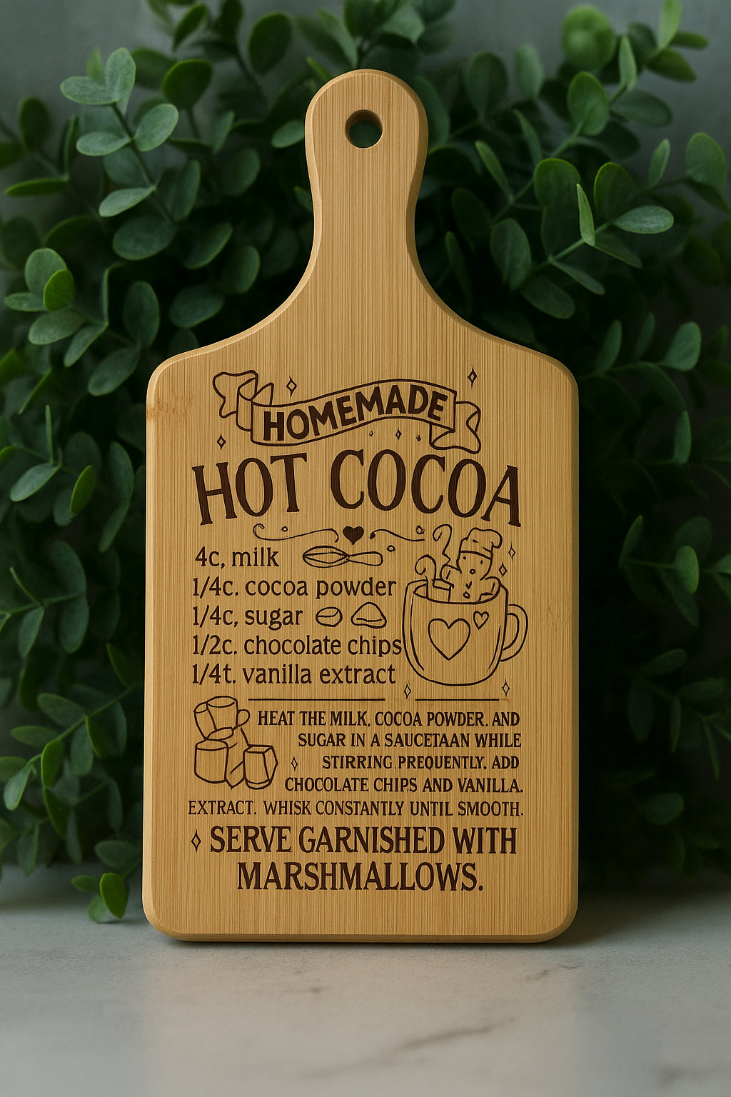 A wooden cutting board engraved with a homemade hot cocoa recipe, surrounded by green leaves. The recipe includes ingredients and instructions, with a drawing of a mug topped with marshmallows and a heart.