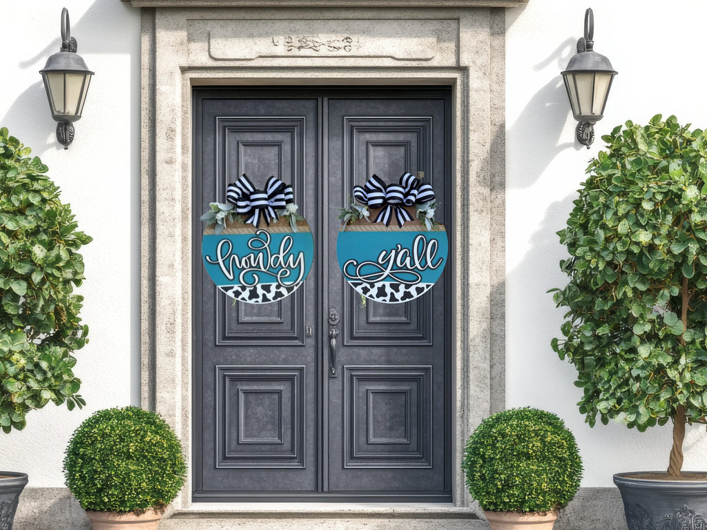 The Howdy Yall Classy Double Door Sign Set features blue and black door wreaths that say “howdy” and “y’all,” framed by stone trim, lantern lights, and potted topiary plants for a warm southern welcome.