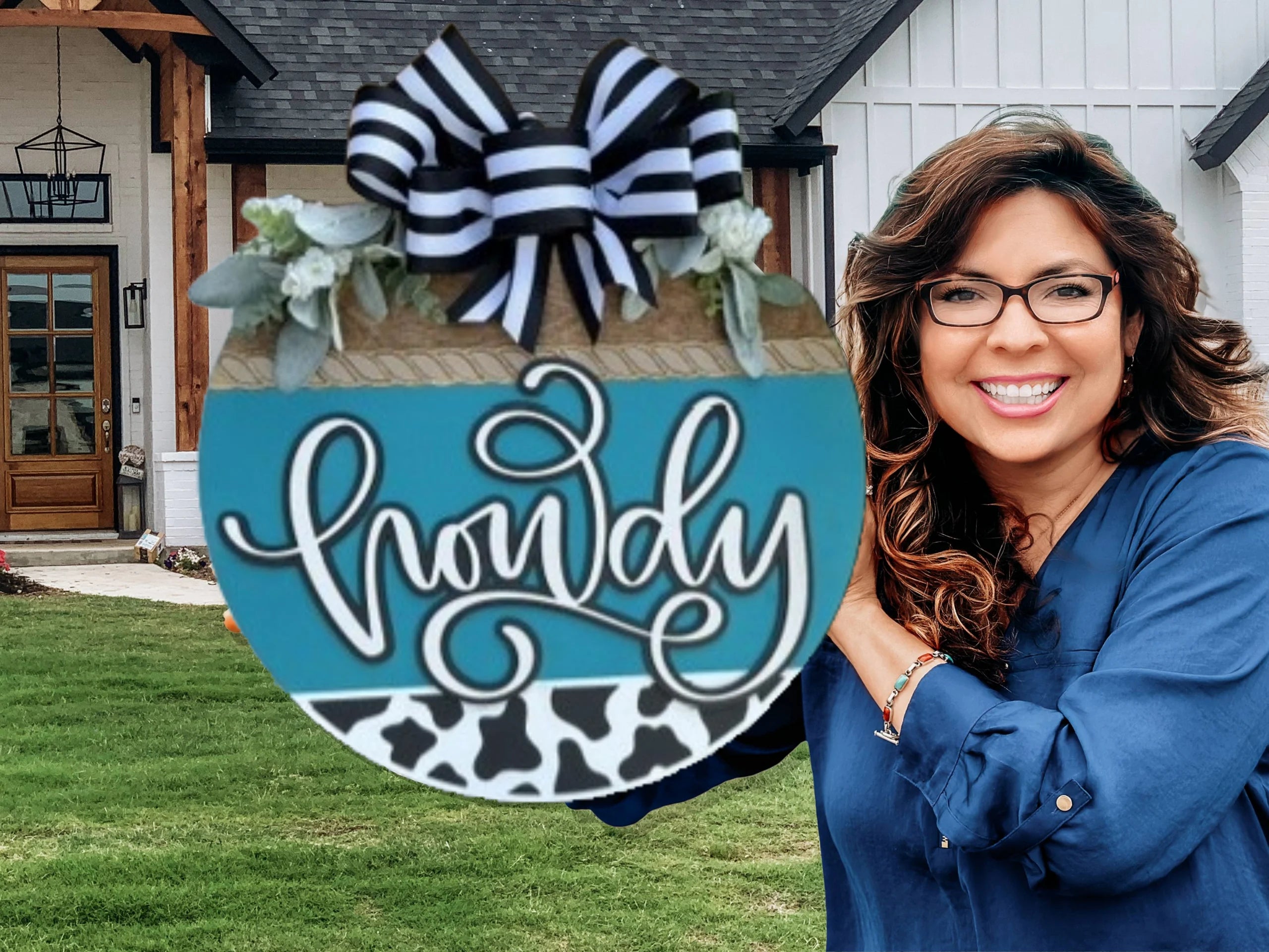A smiling woman with long brown hair and glasses stands outside a house, holding a round blue sign decorated with a striped bow, leaves, and the word howdy in cursive above a black and white cow print strip.