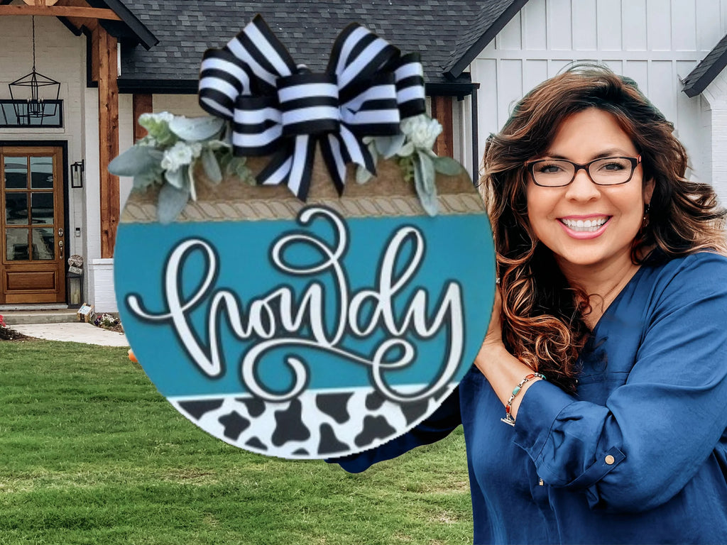 A smiling woman with long brown hair and glasses stands outside a house, holding a round blue sign decorated with a striped bow, leaves, and the word howdy in cursive above a black and white cow print strip.