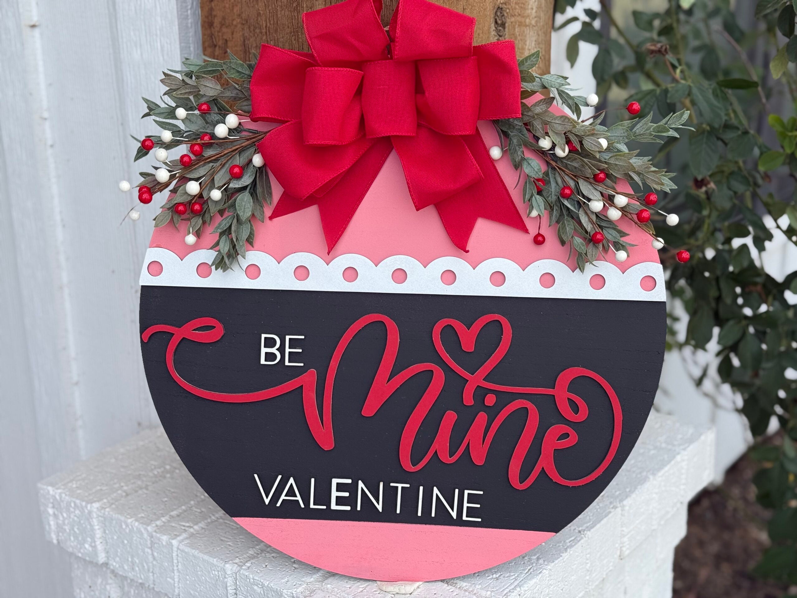 A round wooden decoration with Be Mine Valentine written in red and white cursive. Adorned with a large red bow and greenery with white and red berries on the top. The background is divided into black and pink sections with a white scalloped border.