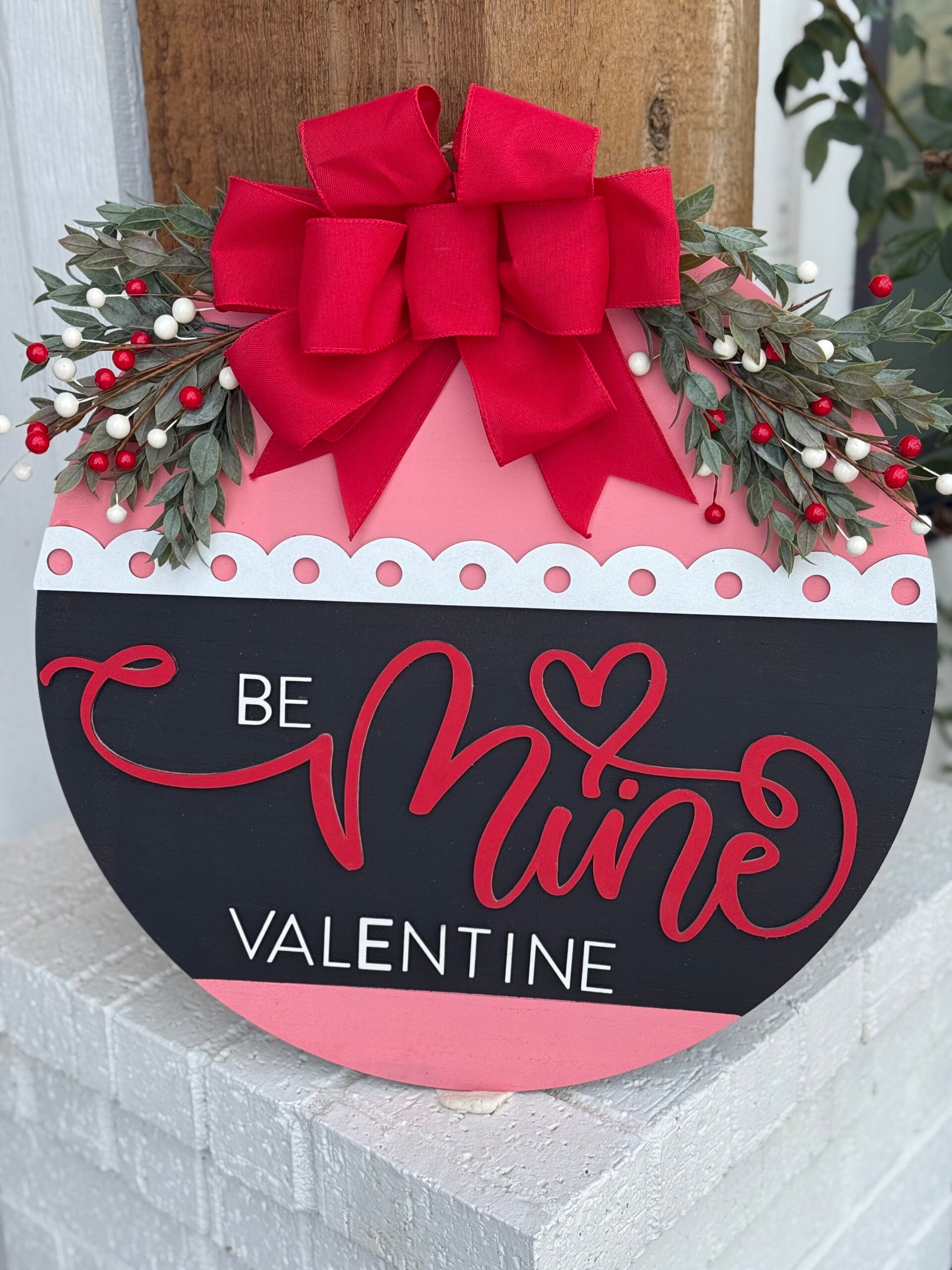 A round Valentine’s Day sign features the words Be Mine Valentine in red and white on a black background. Its decorated with a large red bow and greenery, accented with small white and red berries, and has a pink and white scalloped border on top.