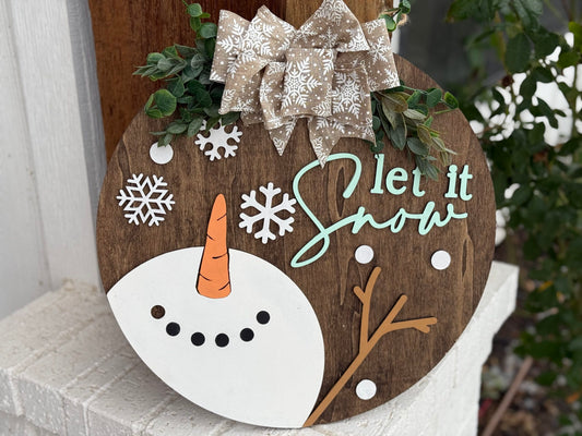 Round wooden sign with Let it Snow written in light blue and white cursive. Decorated with a partial snowman face, white snowflakes, and snow dots. A beige fabric bow with white snowflakes is at the top. Greenery accents are attached around the bow.