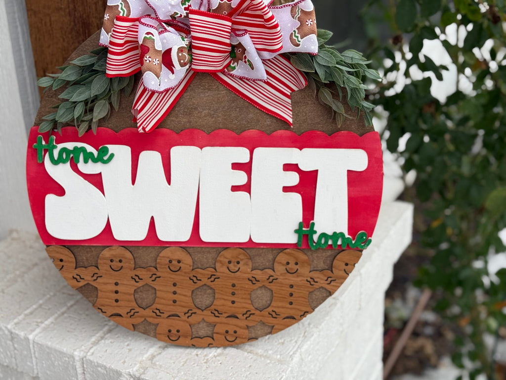 A circular wooden sign adorned with greenery and a festive bow. The sign reads Home SWEET Home in white and green letters on a red background. The bottom half features smiling gingerbread figures. A red and white striped bow with gingerbread patterns is at the top.