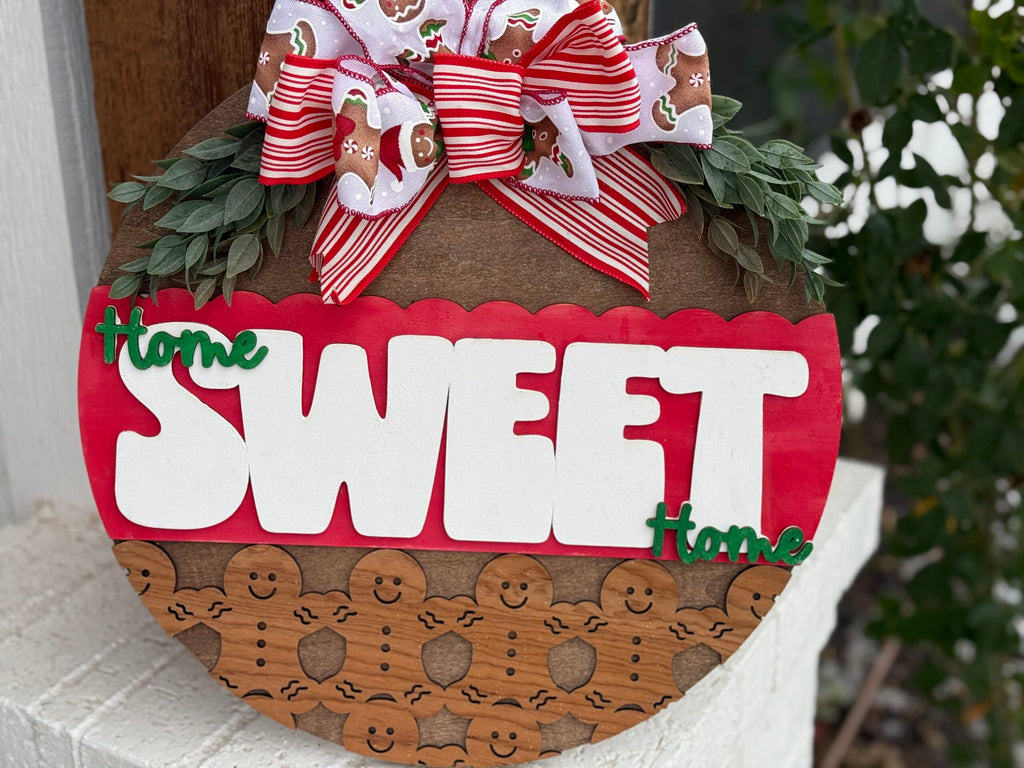Wooden round plaque with Home Sweet Home in large white and green letters. The background features gingerbread men and a red scalloped design. Topped with a festive bow adorned with gingerbread figures and red stripes. Leaves decorate the top edge.